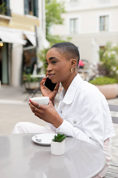 Stylish african american woman talking on cellphone and holding cup of coffee in outdoor cafe in Treviso - Photo, Image