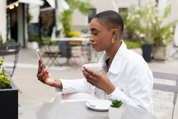 Stylish african american woman holding cup of cappuccino and using smartphone on urban street in Treviso - Zdjęcie, obraz