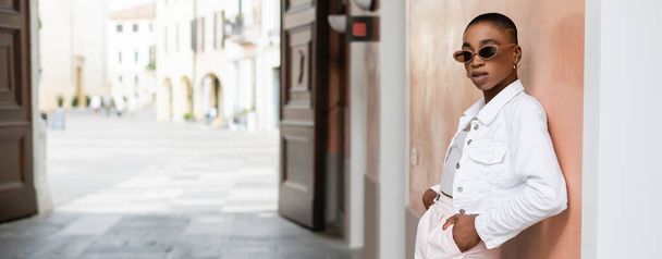 Mujer afroamericana de pelo corto en gafas de sol de pie cerca del edificio en la calle en Treviso, pancarta  - Foto, Imagen