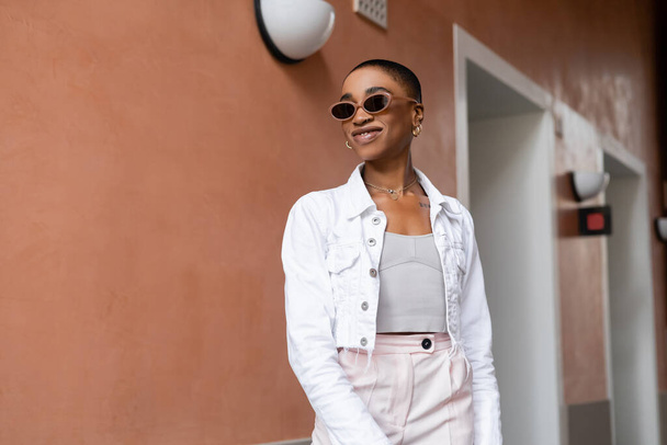 Uśmiechnięta Afroamerykanka w okularach przeciwsłonecznych i białej kurtce stojąca na miejskiej ulicy  - Zdjęcie, obraz
