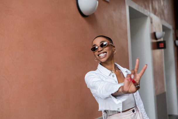Cheerful and stylish african american woman showing peace sign on urban street  - Zdjęcie, obraz