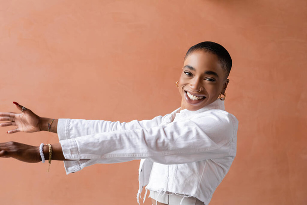 Χαμογελαστή και κομψή Αφροαμερικανή γυναίκα απλώνει τα χέρια και κοιτάζει την κάμερα σε εξωτερικούς χώρους  - Φωτογραφία, εικόνα