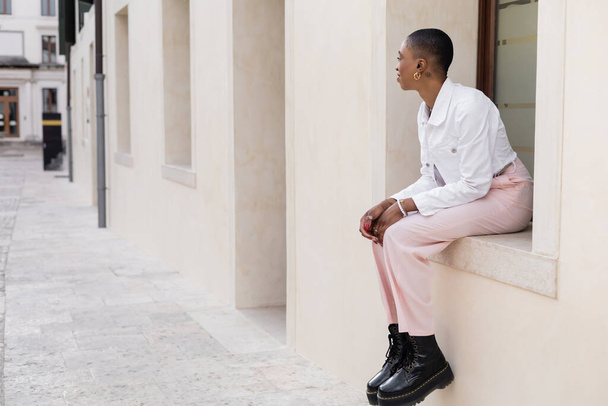 Side view of stylish african american woman sitting on windowsill of building on city street in Treviso - Foto, Bild