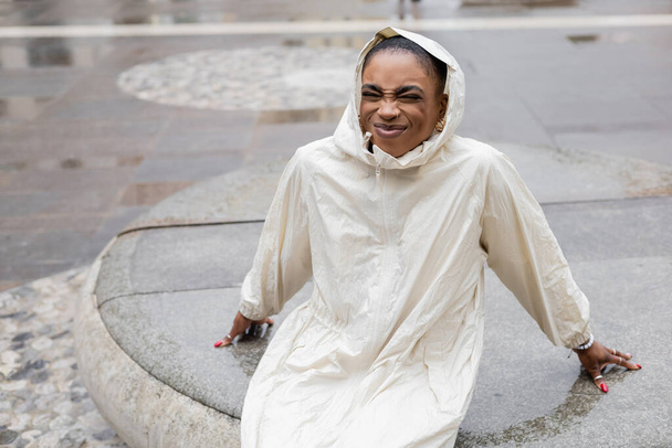 Cheerful african american woman in raincoat sitting on urban street in Treviso - Photo, Image
