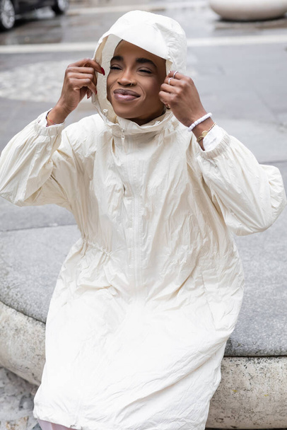 Giovane donna afroamericana in impermeabile seduta su strada urbana offuscata  - Foto, immagini