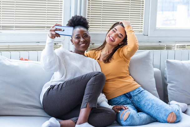 Twee mooie jonge vrouwen nemen selfie op zonnige dag thuis. Vriendinnen. Communicatie en roddels. Houden van Kaukasische en Afro-Amerikaanse meisjes uiten positieve emoties om camera. - Foto, afbeelding
