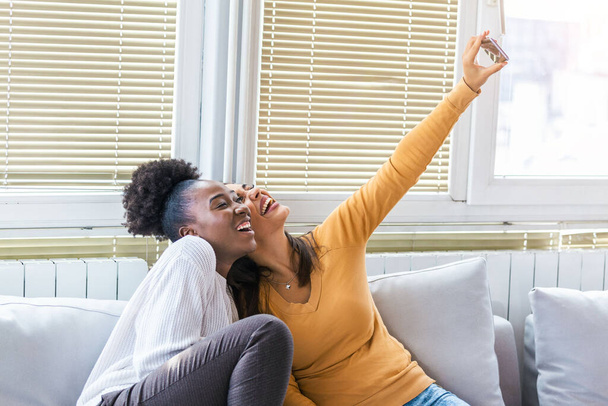 Twee mooie jonge vrouwen nemen selfie op zonnige dag thuis. Vriendinnen. Communicatie en roddels. Houden van Kaukasische en Afro-Amerikaanse meisjes uiten positieve emoties om camera. - Foto, afbeelding