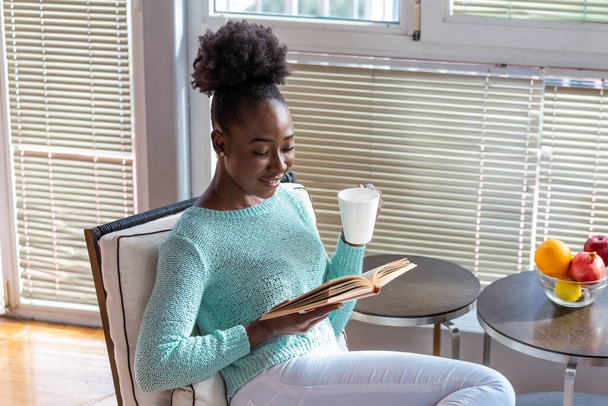 Elinde kitap ve bir fincan sıcak içecekle genç bir kadın. Kitap okuyan güzel bir Afro-Amerikalı kız kütüphanede oturuyor ve arkada kitap rafları var ve kahvesinin tadını çıkarıyor.. - Fotoğraf, Görsel