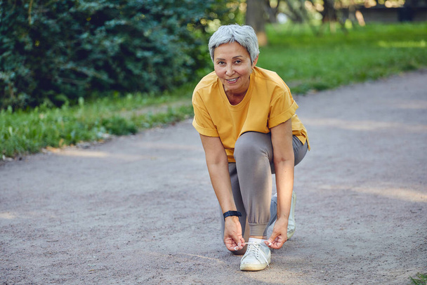 Elderly middle-aged Asia woman in sportive wear ties shoelaces ready for morning jogging or sportive walk stroll in summer park smile look at camera. Healthy active lifestyle of retiree, work out - Photo, image