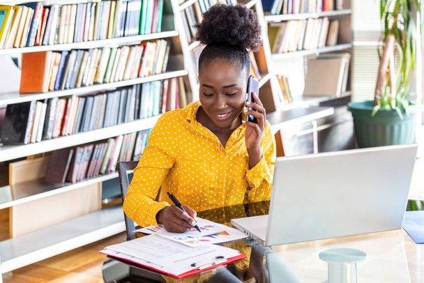 Kobieta sukcesu pracująca w biurze, Afroamerykanka rozmawiająca przez telefon komórkowy i pracująca na laptopie. Koncepcja małych przedsiębiorstw - Zdjęcie, obraz