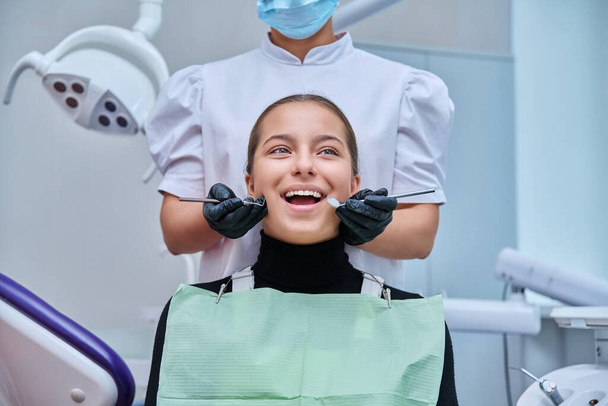 Portrait of young teenage girl in dental chair with hands of doctor with tools. Female teenager smiling with teeth looking at camera in dentist office. Adolescence hygiene treatment dental health care - Foto, afbeelding