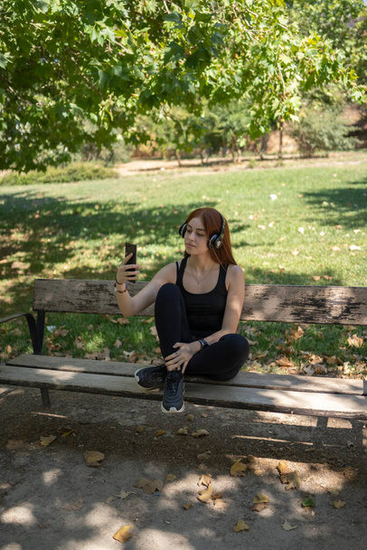 giovane ragazza rossa, guardando lo smartphone, con cuffie musicali sedute su una panchina del parco - Foto, immagini