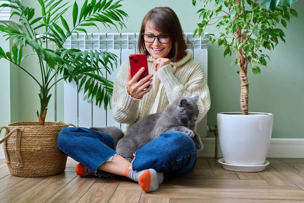 Middle aged woman resting at home in warm clothes using smartphone, warming herself with pet cat near home heating radiator. Winter autumn heating season, lifestyle leisure save energy, crisis concept - Foto, imagen