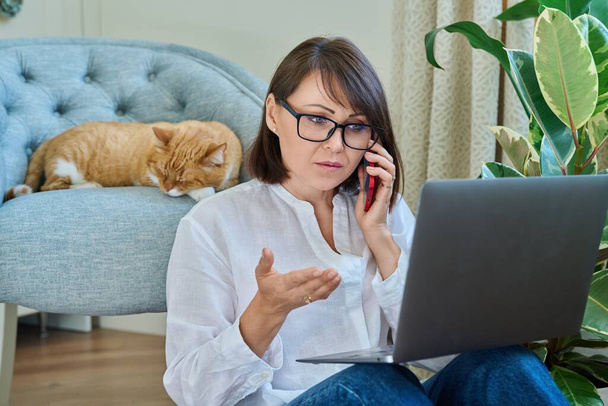 Middle aged woman sitting at home with laptop, working remotely with pet red ginger cat sleeping on armchair. Female freelancer using smartphone laptop, home interior. Technology work people animal - Valokuva, kuva