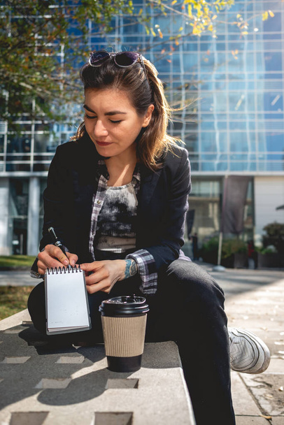 Young, successful and concentrated latina businesswoman with sunglasses and a cup of coffee taking notes of their ideas in a notebook in a park between skyscrapers - 写真・画像