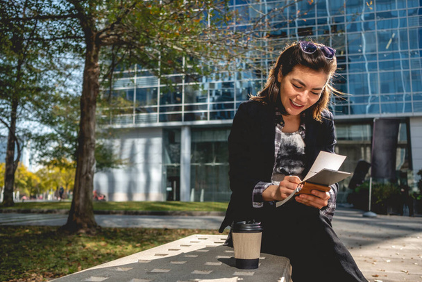 Jovem, bem sucedida e feliz mulher de negócios latina com óculos de sol e uma xícara de café tomando notas de suas ideias em um caderno em um parque entre arranha-céus - Foto, Imagem