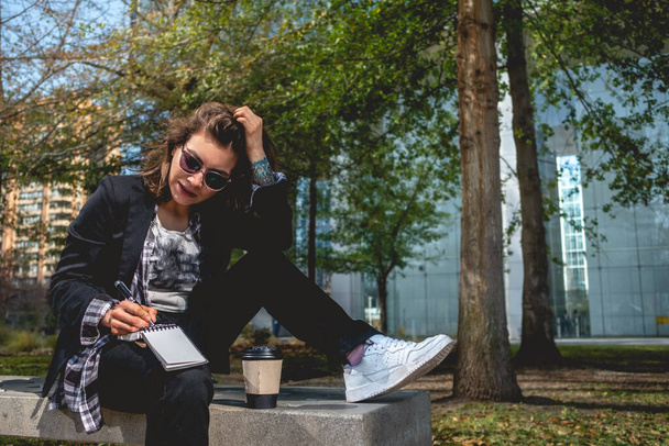 Молода, успішна і концентрована латиноамериканська бізнес-леді з сонцезахисними окулярами і чашкою кави, яка записує свої ідеї в блокноті в парку між хмарочосами
 - Фото, зображення