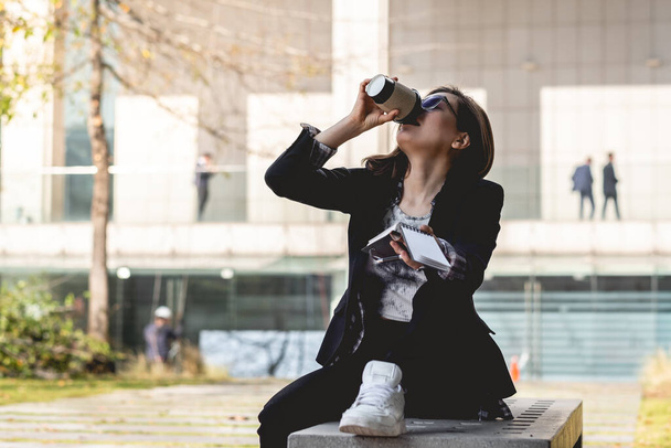 Young, successful and happy latina businesswoman with sunglasses and white sneakers drinking a cup of coffee and taking notes of their ideas in a notebook in a square between skyscrapers - Фото, зображення