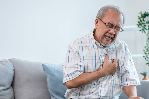 An agonizing senior man suffering from chest pain or heart attack alone in his living room. Serious health problem and feeling unwell concept for seniors. - Foto, Imagen