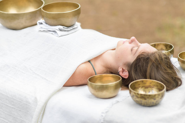 Young woman laying on a massage bed with tibetan singing bowl outdoor. Nepal copper singing bowl sound massage therapy is Buddhist healing practices. Caucasian woman relax with alternative medicine - Photo, Image