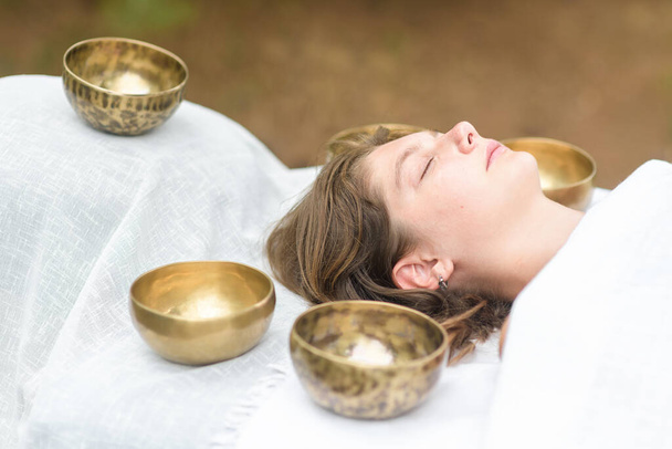 Young woman laying on a massage bed with tibetan singing bowl outdoor. Nepal copper singing bowl sound massage therapy is Buddhist healing practices. Caucasian woman relax with alternative medicine - Photo, Image