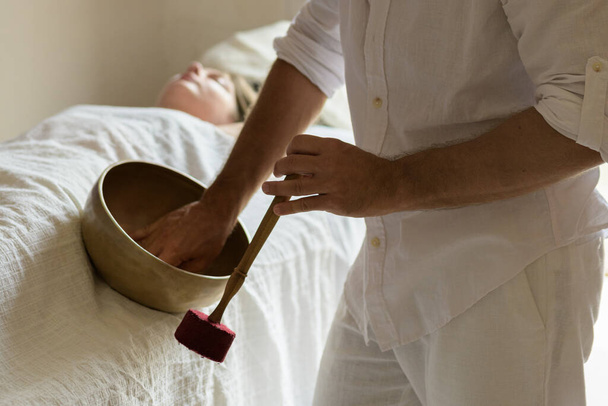 Young woman laying on a massage bed with tibetan singing bowl outdoor. A master of sound massage therapy doing a Buddhist healing practices, male hand holding a felt clapper - Photo, Image