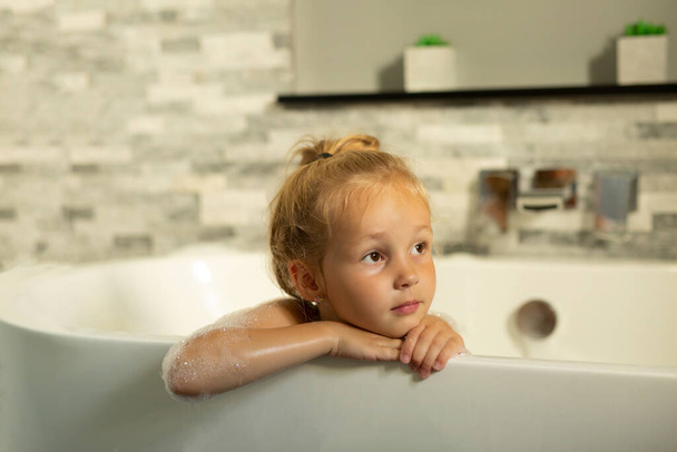 A cute little girl is bathing in a bathtub with white foam. The concept of cleanliness, hygiene and childhood. - Фото, изображение