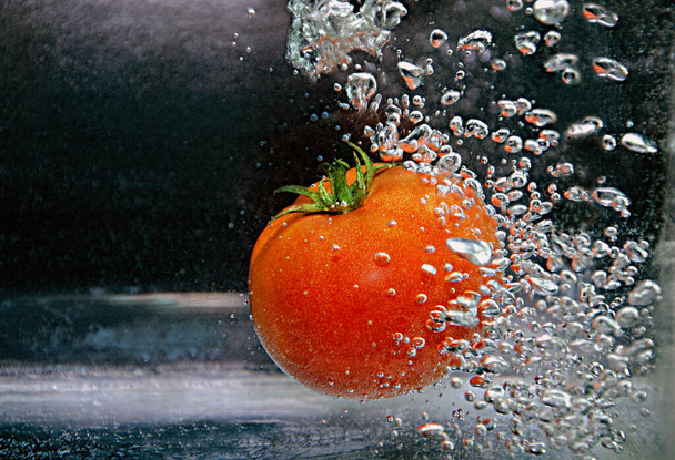 ντομάτα πέφτει σε καθαρό νερό με μια μεγάλη βουτιά σε σκούρο φόντο. ντομάτα πέφτει στο νερό με πιτσιλιές και φυσαλίδες - Φωτογραφία, εικόνα