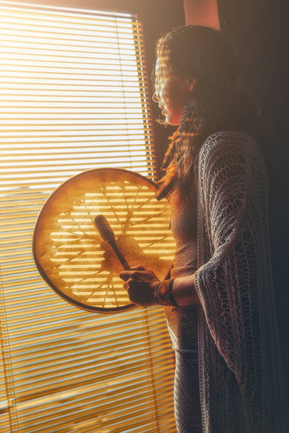 красивая шаманская девушка играет на барабане шамана в доме у окна - Фото, изображение