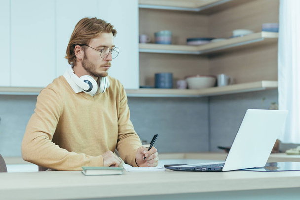 Young serious and focused man studying remotely at home, student in kitchen wearing glasses using laptop for distance learning taking notes on study material. - Фото, зображення