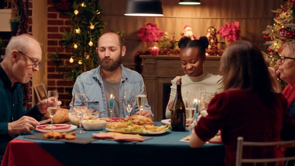 Χαρούμενοι πολυπολιτισμικοί άνθρωποι που απολαμβάνουν το Χριστουγεννιάτικο δείπνο τρώγοντας ψητό κοτόπουλο. Ευτυχισμένα και ποικιλόμορφα μέλη της οικογένειας απολαμβάνοντας παραδοσιακά σπιτικά μαγειρεμένα τρόφιμα Wihle γιορτάζει τις χειμερινές διακοπές μαζί. - Φωτογραφία, εικόνα