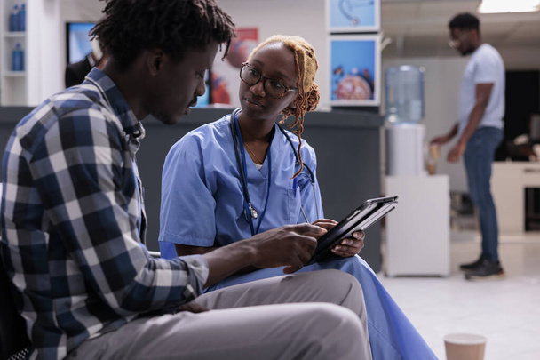 Afro-Amerikalı uzman ve hastalıktan bahseden bir adam, hastanenin bekleme salonunda hasta bir adamla check-up yapıyor. Tıbbi yardım uzmanı ilaç verecek kişi, sağlık merkezi. - Fotoğraf, Görsel
