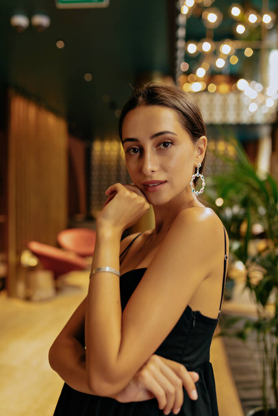 Alluring young caucasian woman in elegant black dress posing at camera against lights in elegant hotel. Femininity and beauty sensuality concept. - Foto, immagini