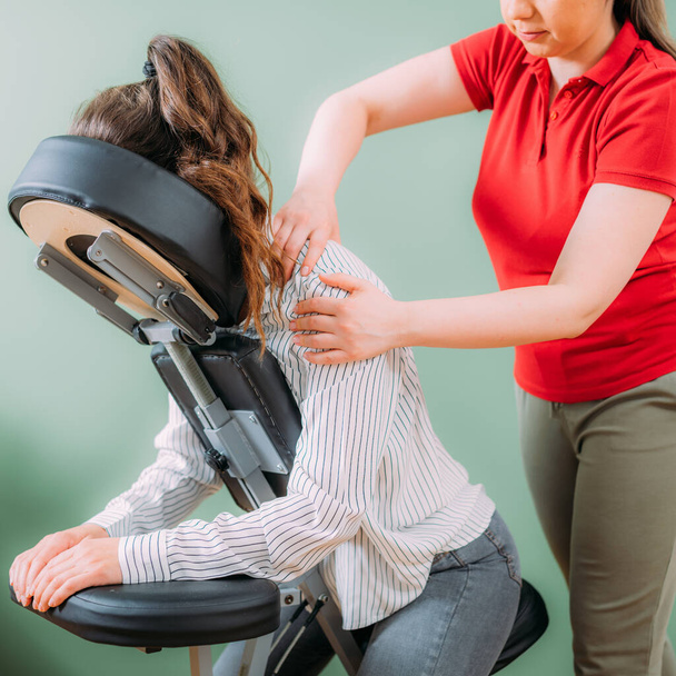 Female employee sitting on a portable massage chair in business office. Therapist massaging her shoulders, releasing muscle tension. - Zdjęcie, obraz