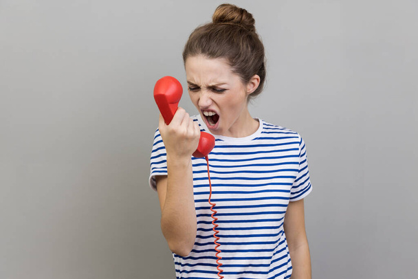 Portrait of angry nervous woman wearing striped T-shirt screaming and yelling talking retro landline phone, complaining on connection quality. Indoor studio shot isolated on gray background. - Фото, изображение