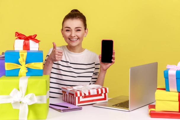 Kind woman sitting at workplace with gift boxes, holding mobile phone with blank display, showing thumb up, mockup screen on mobile device. Indoor studio studio shot isolated on yellow background. - Фото, зображення