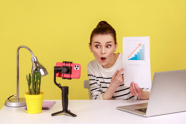 Astonished woman employee analyzing graph of clients growth, recording video on smartphone camera or streaming online, conducting conference. Indoor studio studio shot isolated on yellow background. - Photo, Image