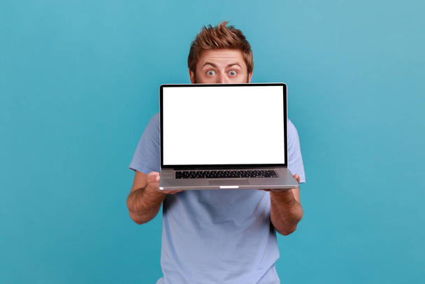 Portrait of man hiding behind white empty screen laptop, surprised with advertising area, looking at camera with big eyes, updating operation system. Indoor studio shot isolated on blue background. - Photo, image