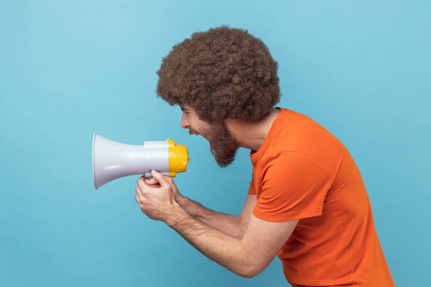 Side view of man with Afro hairstyle wearing orange T-shirt loudly screaming at megaphone, making announce, protesting, wants to be heard. Indoor studio shot isolated on blue background. - Foto, Bild