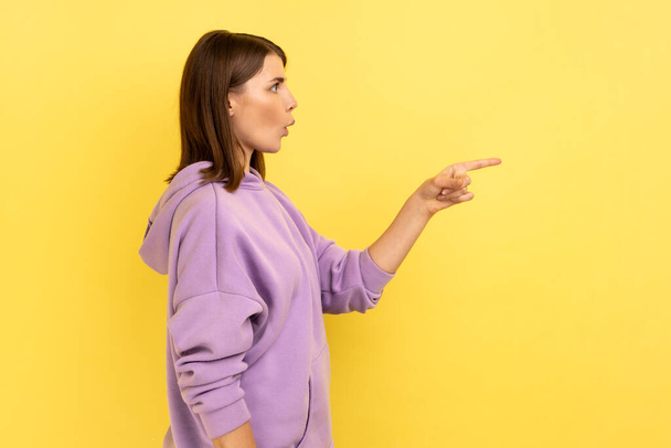 Side view of excited beautiful young adult woman standing and pointing at copyspace with surprised face, wearing purple hoodie. Indoor studio shot isolated on yellow background. - Foto, Imagem