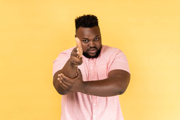 I'll kill you. Portrait of man wearing pink shirt pointing finger guns to camera and looking aggressive, threatening to shoot, hands imitating weapon. Indoor studio shot isolated on yellow background. - Photo, image