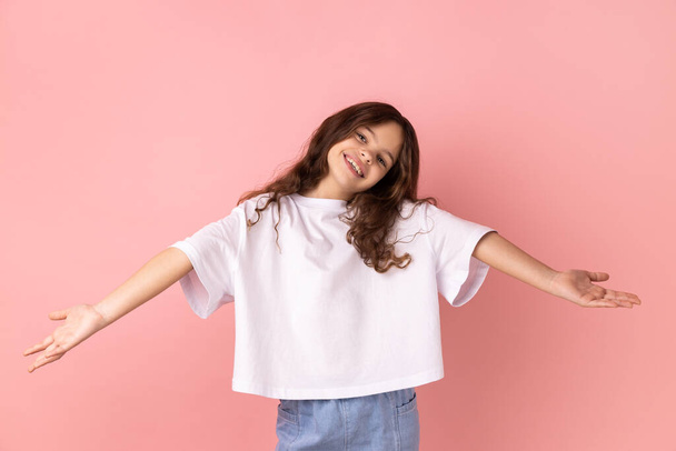 Portrait of positive optimistic little girl wearing white T-shirt inviting to embrace, keeping hands as giving, sharing for free. Indoor studio shot isolated on pink background. - Fotografie, Obrázek
