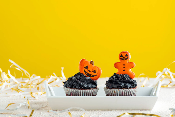 Holiday cupcakes with black cream to celebrate halloween. Muffins decorated with gingerbread on yellow background. - Photo, Image