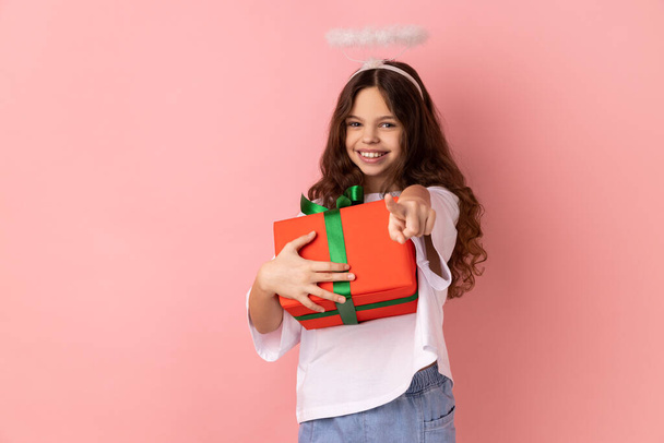 Little girl wearing white T-shirt with holy nimbus with kind smile holding gift box, charity organization, pointing to camera with finger, religion. Indoor studio shot isolated on pink background. - Photo, image