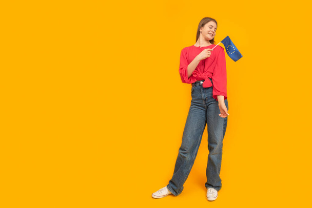 Full-length portrait of young girl with the EU flag in her hands on orange background. Study in Europe. Copy space - Foto, Bild