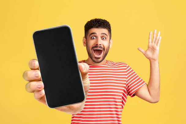 Amazed cheerful man with beard wearing red T-shirt showing mobile phone with blank display, expressing happiness, raised arm and screaming happily. Indoor studio shot isolated on yellow background. - Photo, image