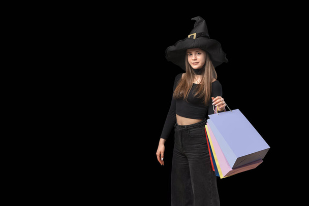 手にカラフルな紙のショッピングバッグと魔女の帽子の若い女性。コピースペース。ブラック・フライデーのコンセプト。黒の背景に隔離された - 写真・画像