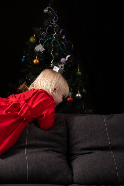 Little girl in red dress on the sofa on christmas tree background - Photo, image