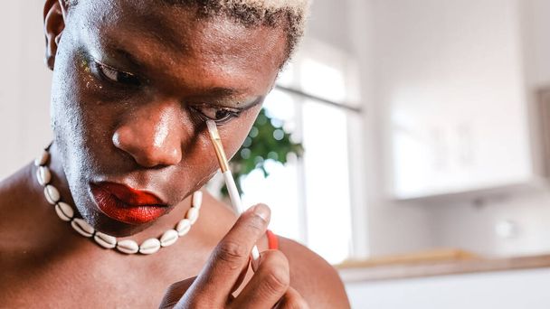 Low angle of African American gay with seashell necklace and red lips drawing line under eye during makeup application in daytime at home - Fotografie, Obrázek