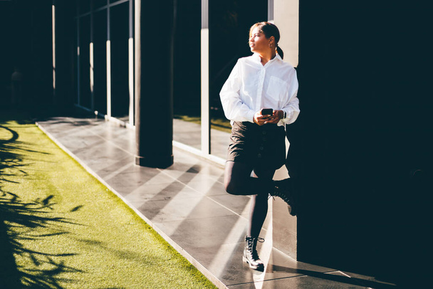 A charming Hispanic plus-size businesswoman is holding a smartphone in her hands while leaning against the wall outside of an office building entrance with stripes of light on the ground from windows - 写真・画像